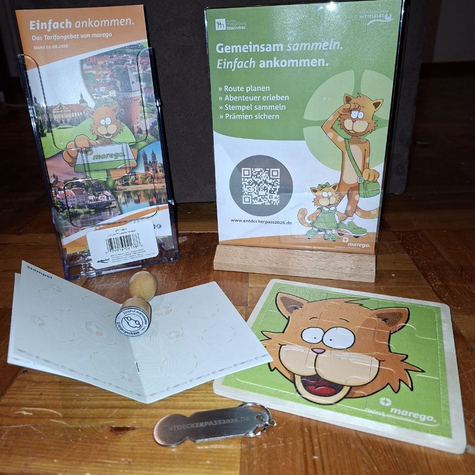 Aufsteller zum Entdeckerpass mit QR-Code, daneben ein Prospekt von marego; davor ein aufgeschlagenes Stempelheft mit Holzstempel, ein Holzpuzzle und ein Einkaufswagenchip mit der Aktionsadresse